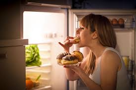 Une femme grignote un dessert sucré devant la porte de son frigo ouverte