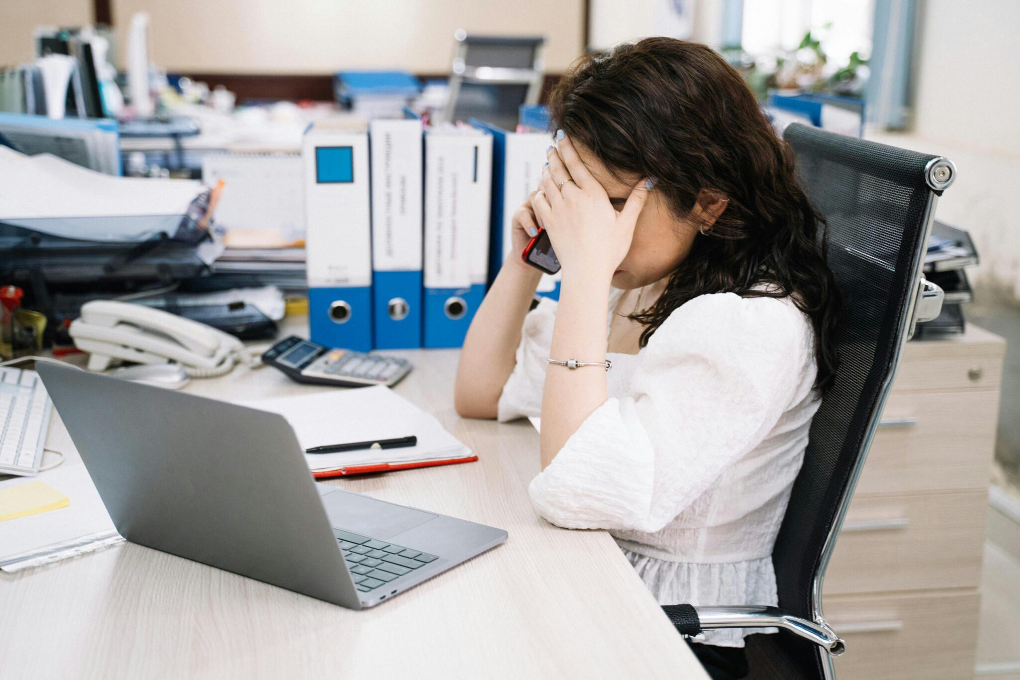 Une femme assise à son bureau, devant son ordinateur et au téléphone, se tenant le front avec sa main, visiblement très stressée et surmenée